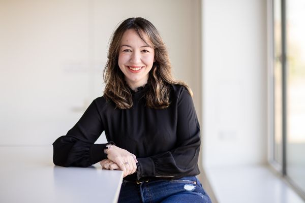 A woman sitting on a stool, leaning on the table with her elbow, smiling at the camera.