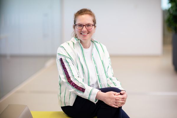 A woman wearing glasses, sitting down with her legs crossed, smiling at the camera