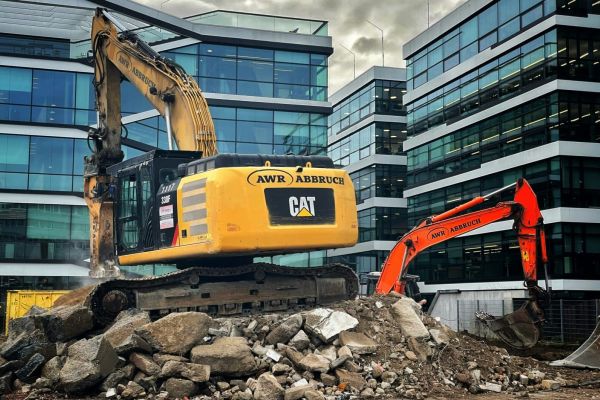 Een gele graafmachine aan het werk bij kantoorgebouwen.
