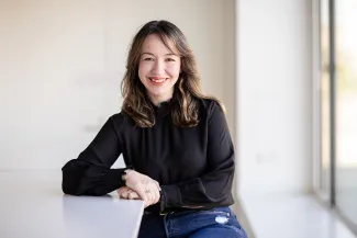 A woman sitting on a stool, leaning on the table with her elbow, smiling at the camera.