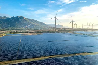 Luchtfoto van een groot zonnepark met honderden zonnepanelen op de voorgrond. Op de achtergrond staan meerdere windturbines, met groene bergen en een heldere blauwe lucht.